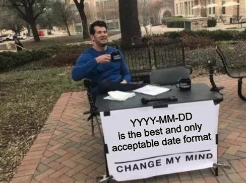 Cheerful man sitting behind a card table outdoors with a cup of coffee. A poster is taped to the front of the table that reads: YYYY-MM-DD is the best and only acceptable date format